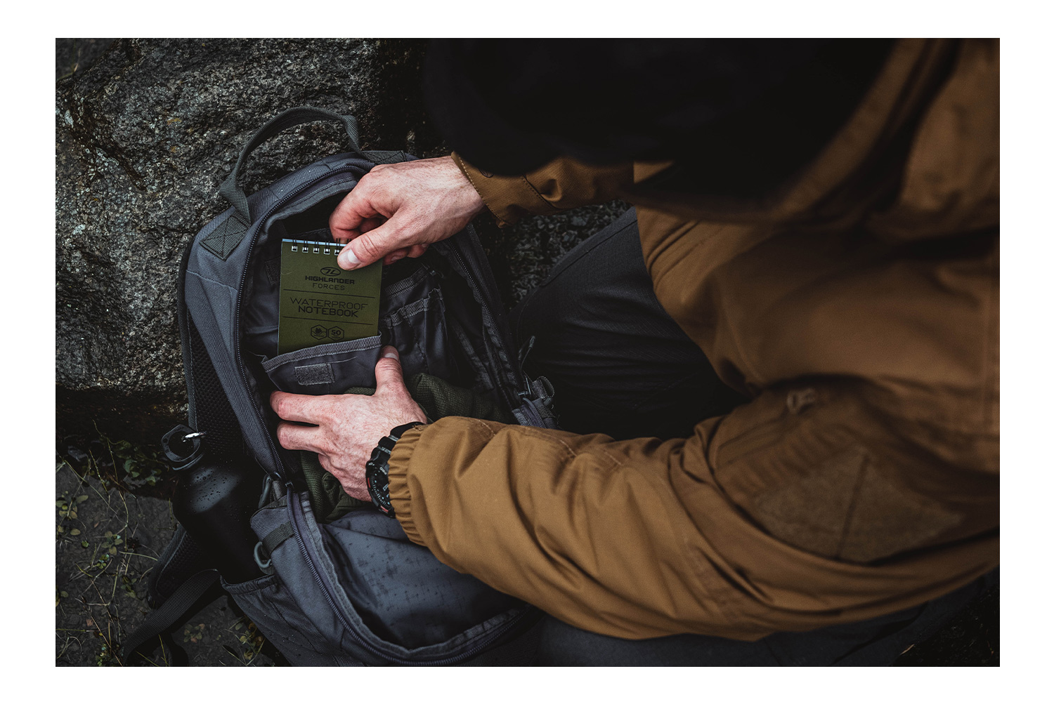 Eine Person in einer braunen Jacke packt draußen ein grünes wasserdichtes Notebook der Highlander Forces in einen grauen taktischen Rucksack.