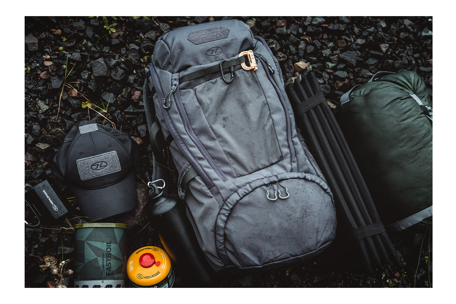 Grauer taktischer Highlander-Rucksack mit Outdoor-Ausrüstung, darunter Mütze, Campingkocher und Schlafsack auf felsigem Boden.