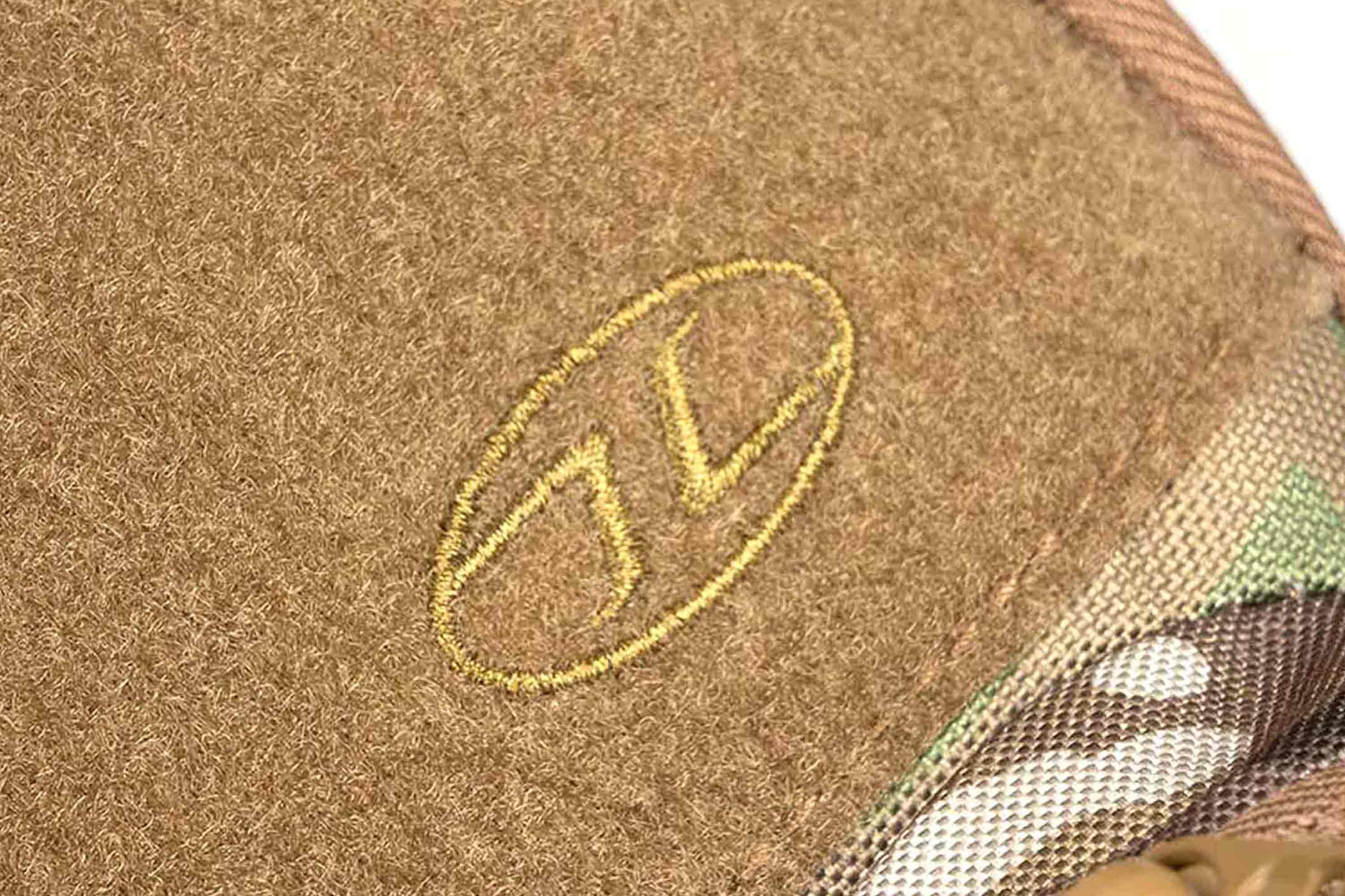 Nahaufnahme eines hellbraunen Aufnähers für taktische Ausrüstung mit einem gestickten goldenen Logo auf einer strukturierten, flauschigen braunen Stoffoberfläche.