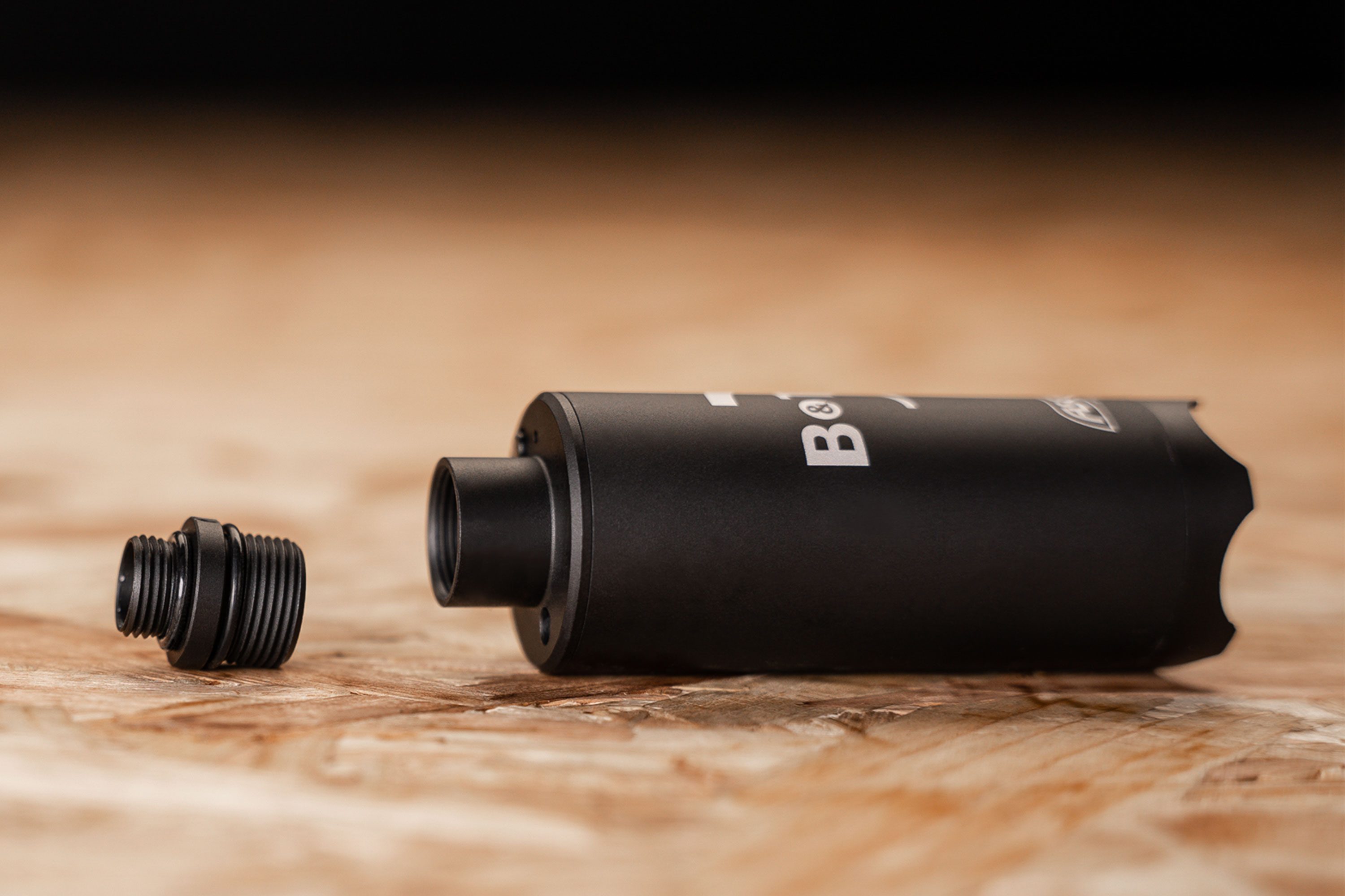 Schwarzer B&T-Schalldämpfer mit abnehmbarem Gewindeadapter, der auf einer Holzoberfläche ruht und für den Einsatz mit Airsoft oder Schusswaffen geeignet ist.