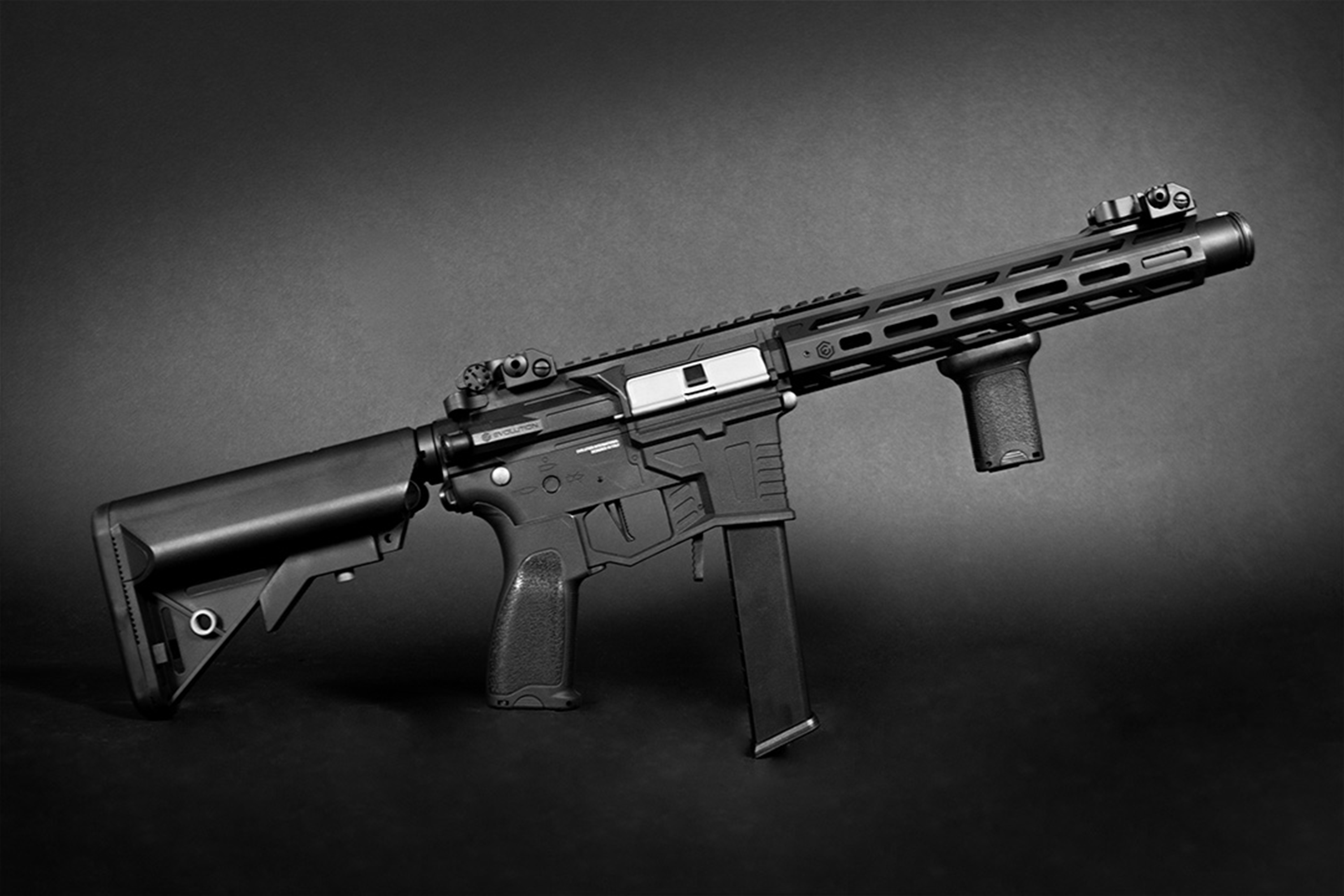 Schwarzer Evolution-Airsoft-Karabiner mit taktischem Handschutz, vertikalem Vordergriff und verstellbarem Schaft auf dunklem Hintergrund.