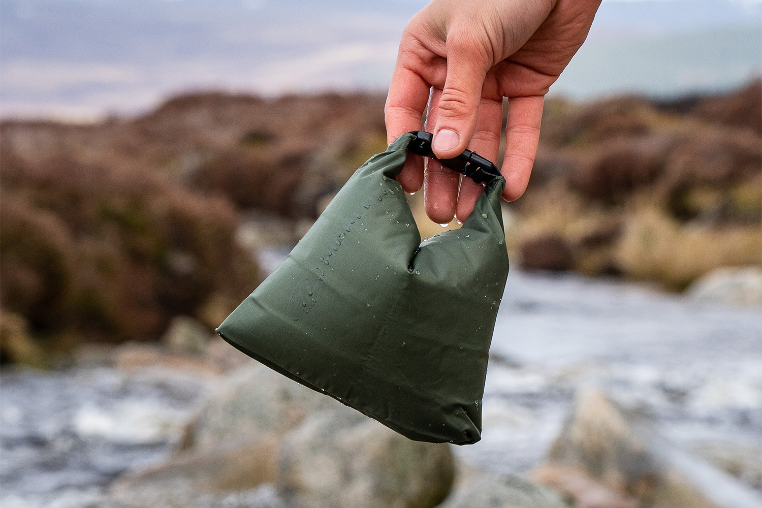 Eine Hand hält einen kleinen, olivgrünen wasserdichten Packsack mit schwarzem Schnallenclip vor einer verschwommenen Außenlandschaft.