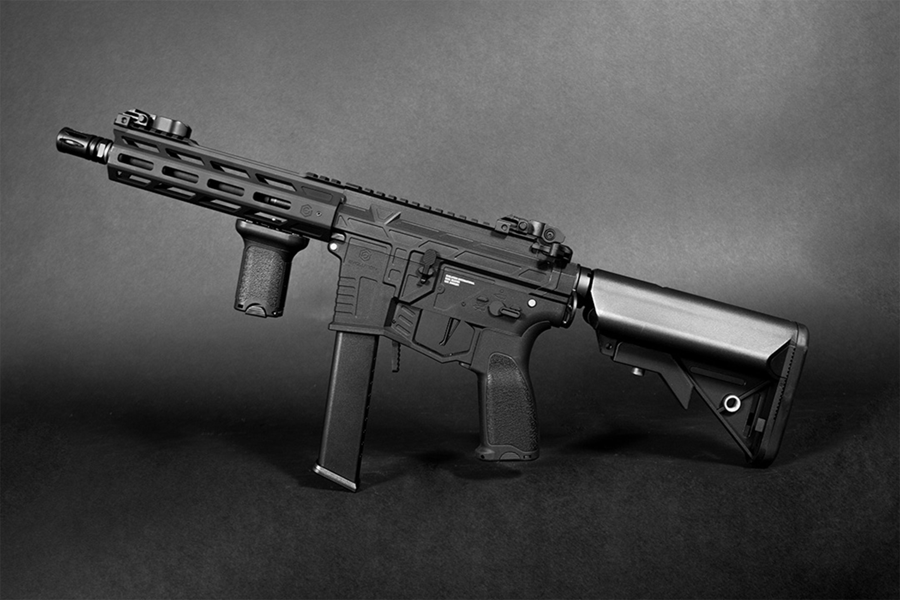Schwarzes Airsoft-Karabinergewehr mit taktischem Handschutz, vertikalem Vordergriff und verstellbarem Schaft auf dunklem Hintergrund.