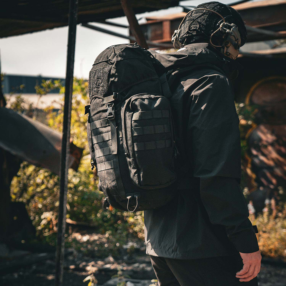 Eine Person, die einen schwarzen taktischen Rucksack mit MOLLE-Gurtband trägt und draußen in einer rauen Industrieumgebung steht.