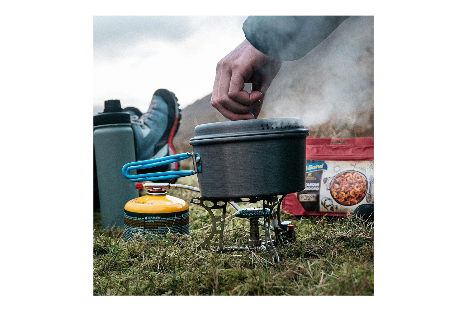 Eine Person kocht mit einem tragbaren Campingkocher und einem Topf auf Gras, in der Nähe befindet sich ein Benzinkanister und ein Essensbeutel im Freien.