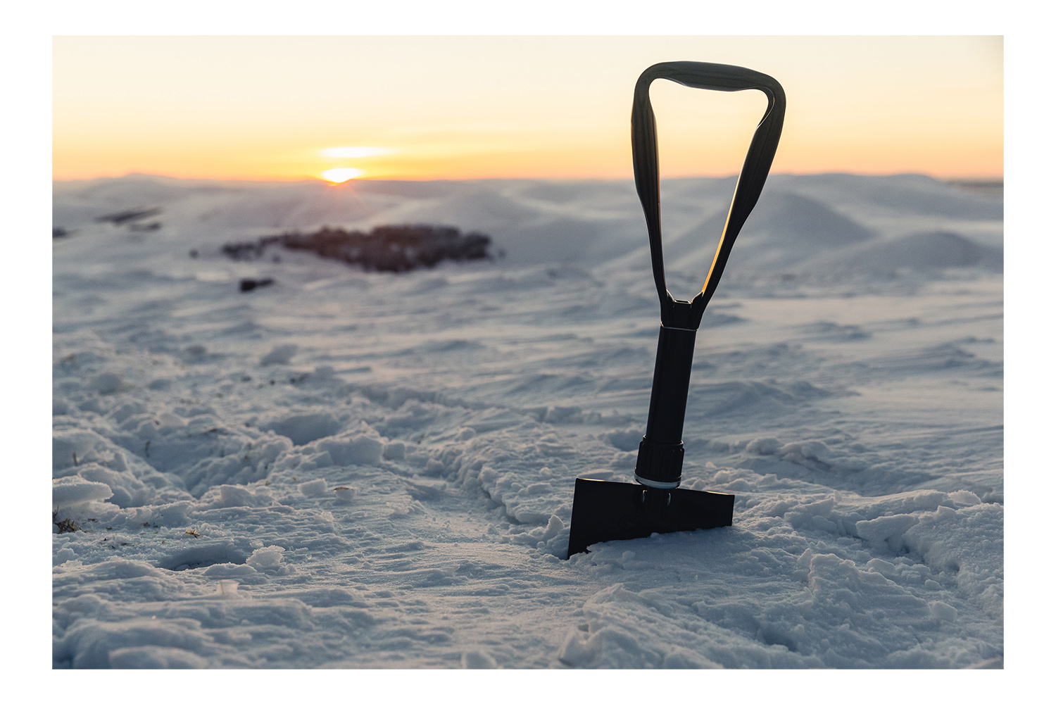 Eine schwarze Klappschaufel steht während eines Sonnenuntergangs aufrecht im tiefen Schnee, ideal für Outdoor-Survival- und Ausrüstungsliebhaber.