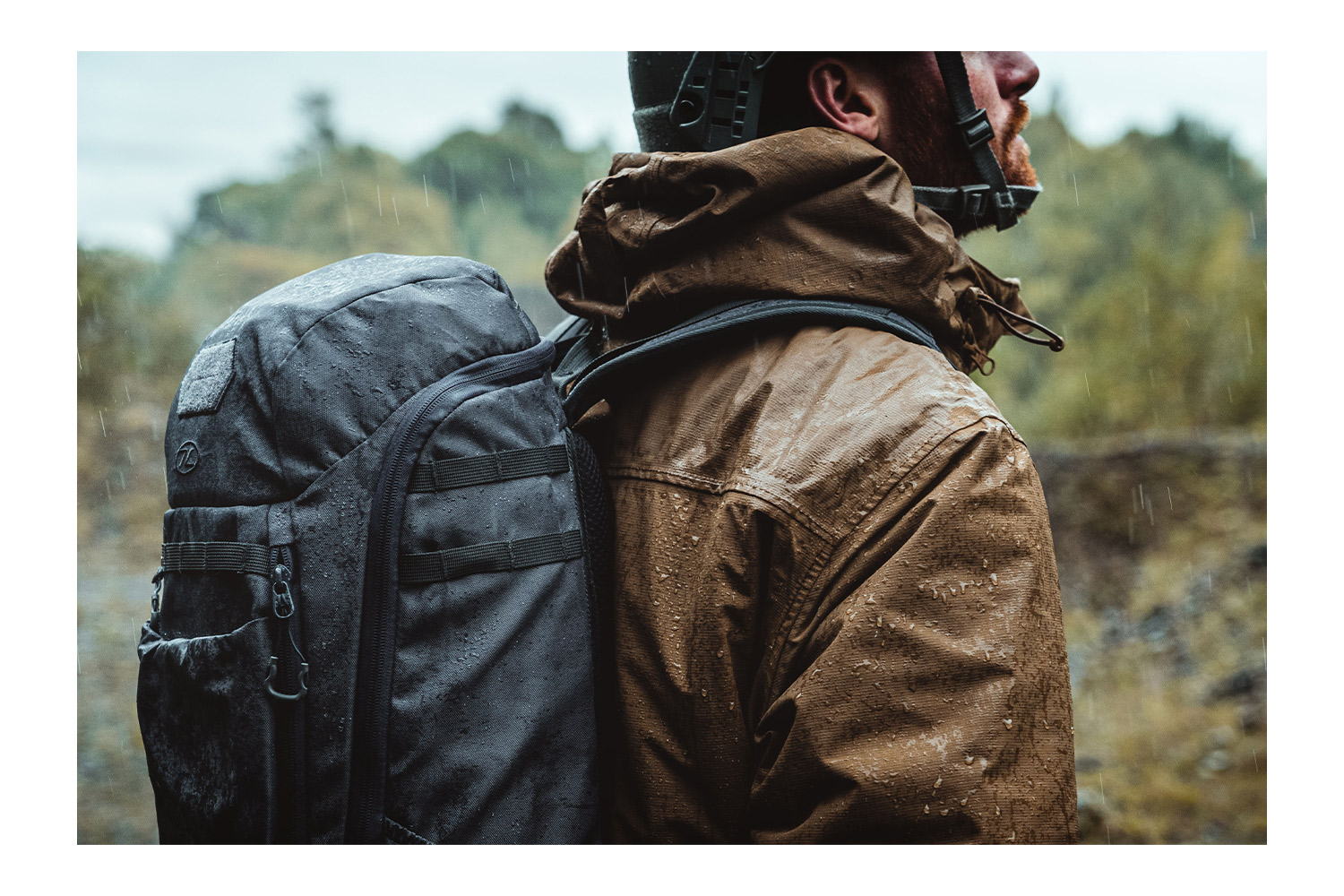 Eine Person trägt eine braune Outdoor-Jacke und einen schwarzen taktischen Rucksack im Regen, aus dem Seitenprofil betrachtet.