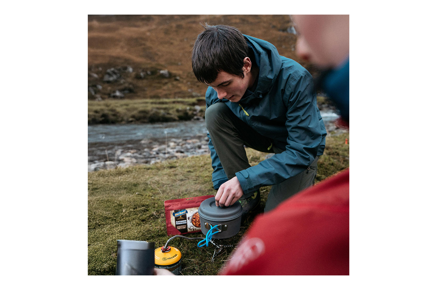 Ein junger Mann in einer blauen Jacke bereitet eine Mahlzeit mit einem tragbaren Campingkocher und Topf in der Nähe eines Flusses in der Wildnis zu.