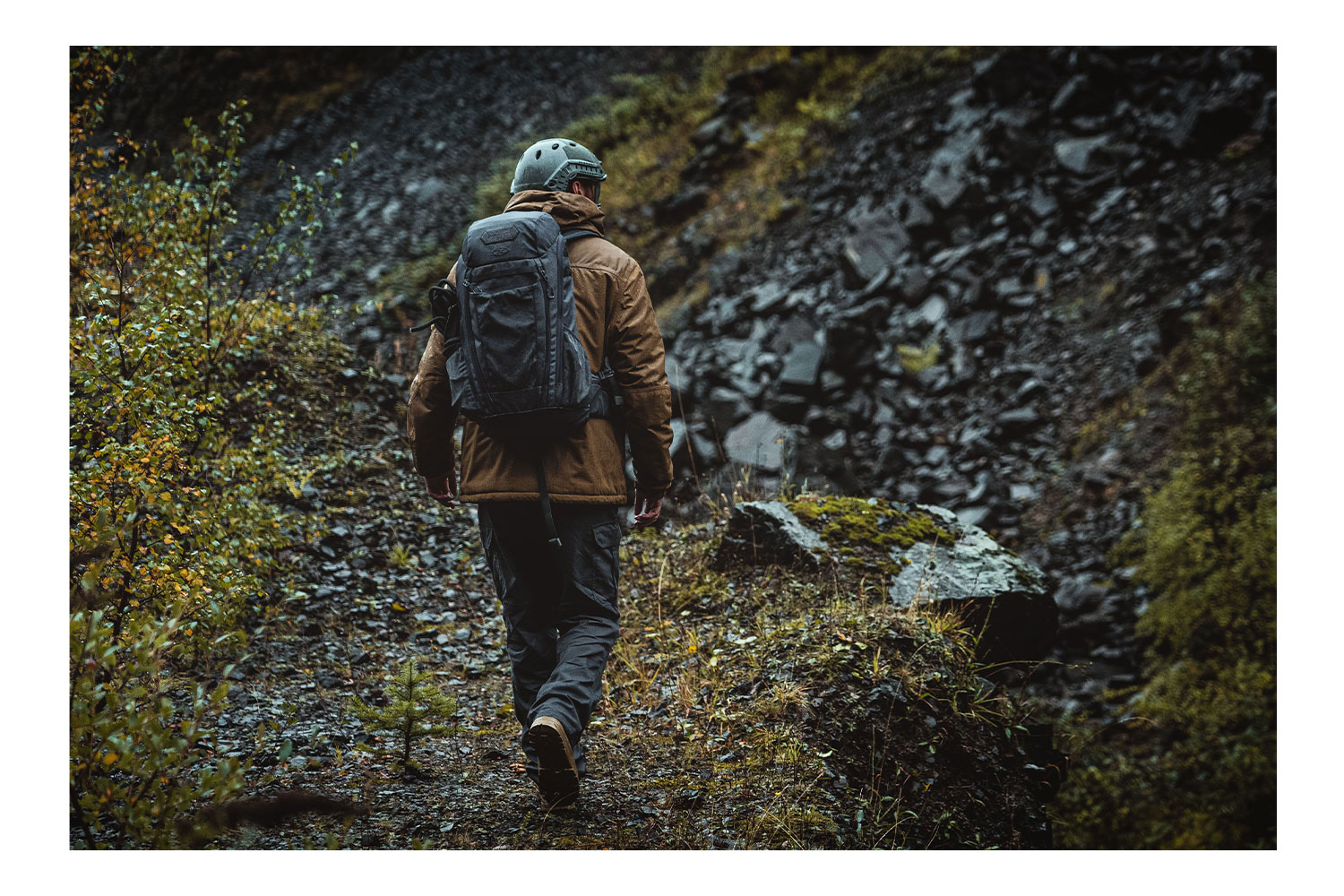 Eine Person mit taktischem Helm, brauner Jacke und grauem Rucksack wandert auf einem steinigen Pfad in der Wildnis.
