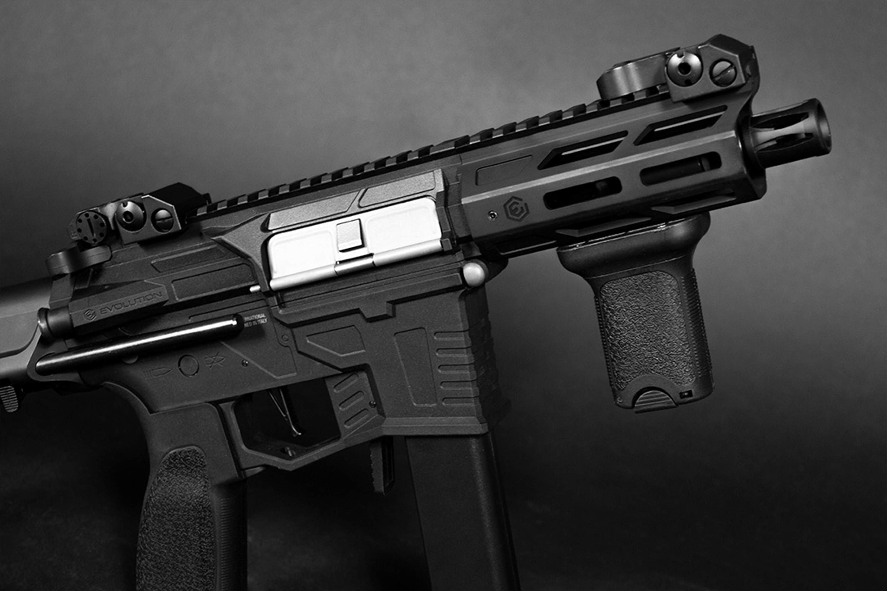 Schwarzer Evolution-Airsoft-Karabiner mit kurzem Lauf, vertikalem Vordergriff und silberner Auswurföffnung auf dunklem Hintergrund.