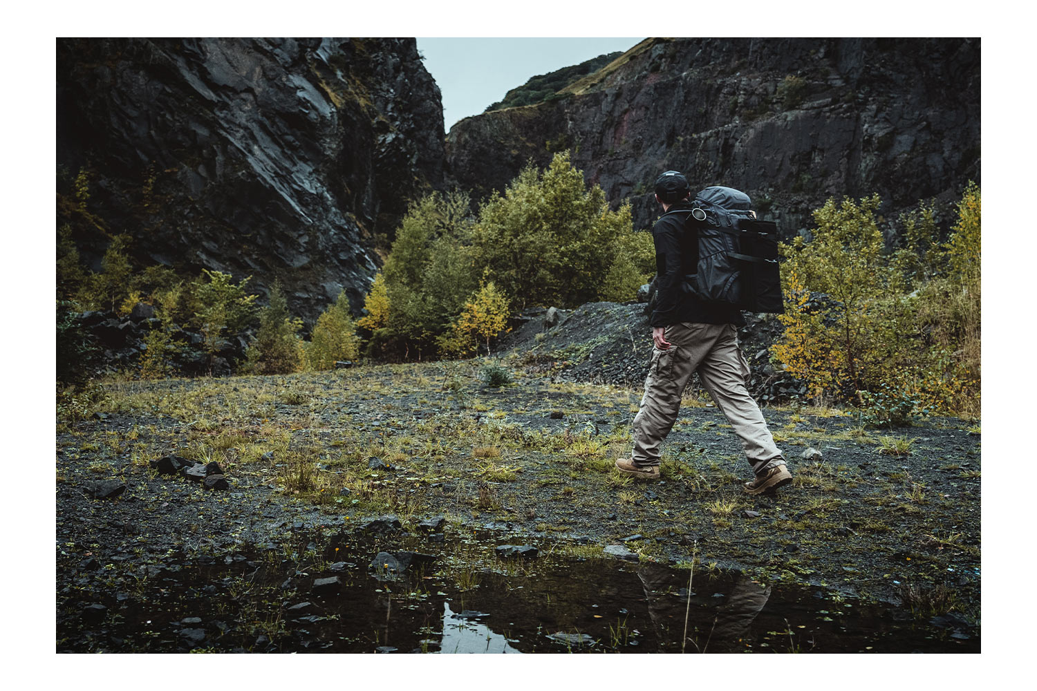 Ein Wanderer mit einem großen taktischen Rucksack wandert durch eine felsige, zerklüftete Landschaft mit Bäumen und steilen Felswänden.