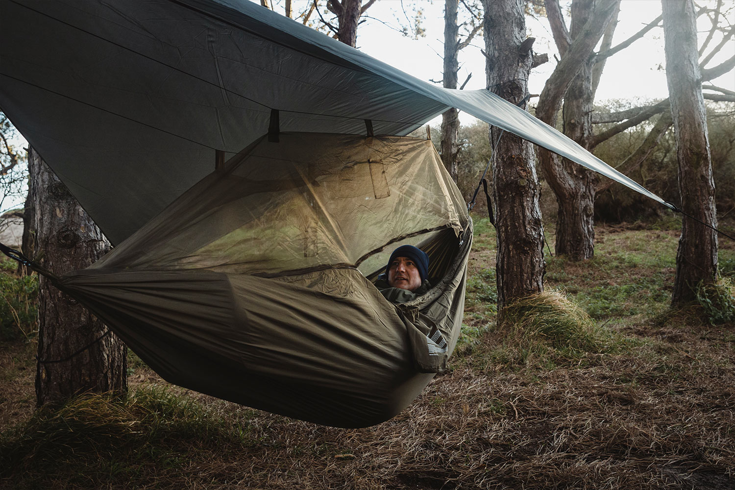 Ein Mann ruht in einer olivgrünen Camping-Hängematte mit Moskitonetz, geschützt durch eine Plane zwischen Bäumen in einem Wald.