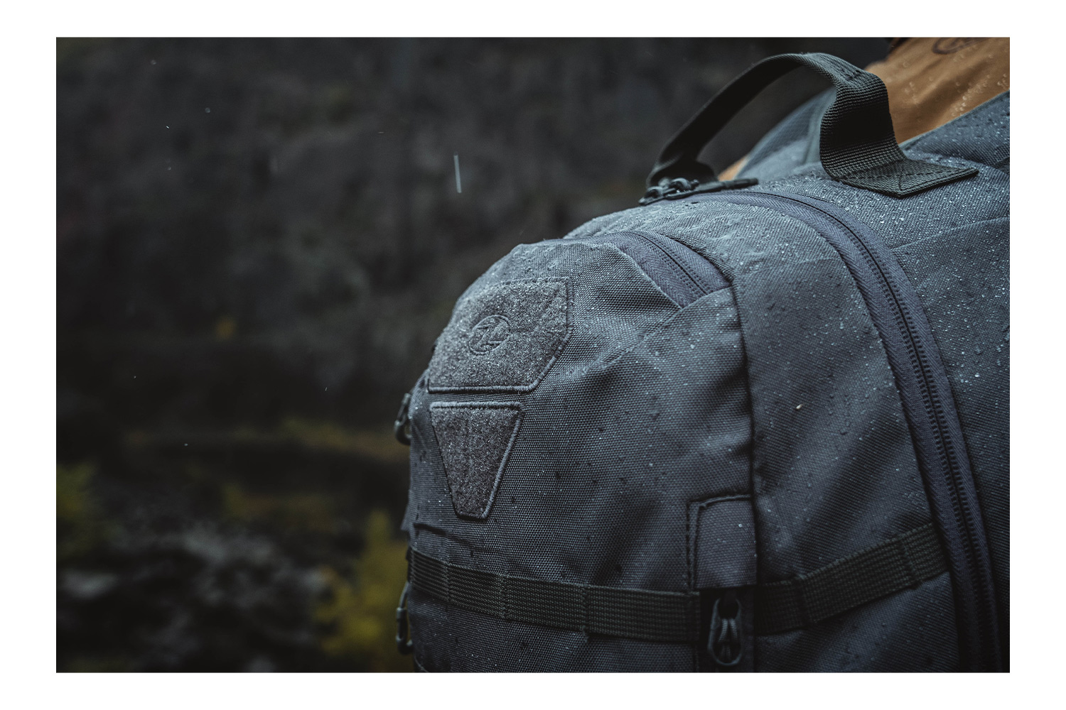 Nahaufnahme eines dunkelgrauen taktischen Rucksacks mit Wassertropfen, mit MOLLE-Gurtband und einem Klettverschlussbereich.