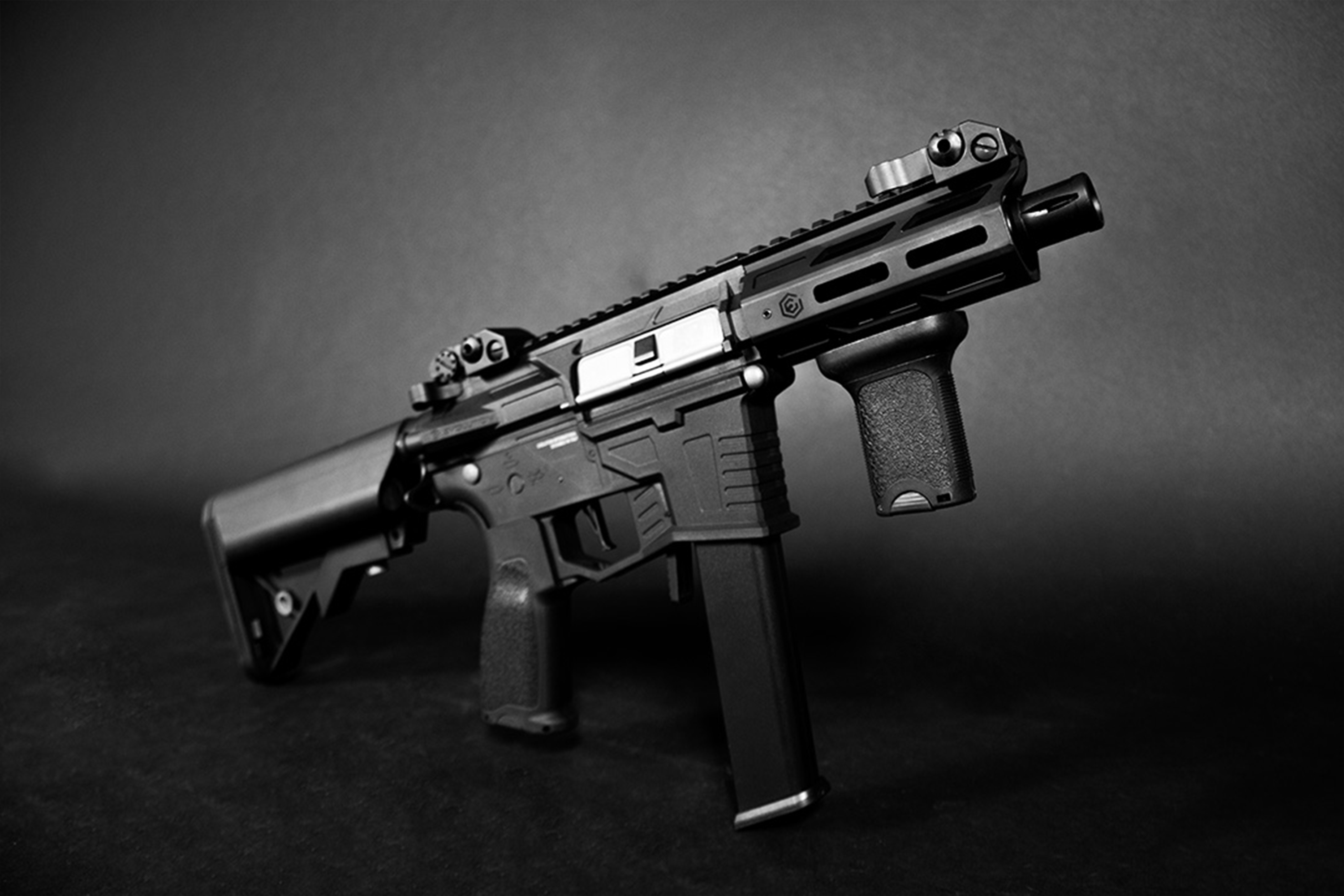Schwarzes taktisches Luftgewehr mit kurzem Lauf, vertikalem Vordergriff und eiserner Visierung vor dunklem Studiohintergrund.
