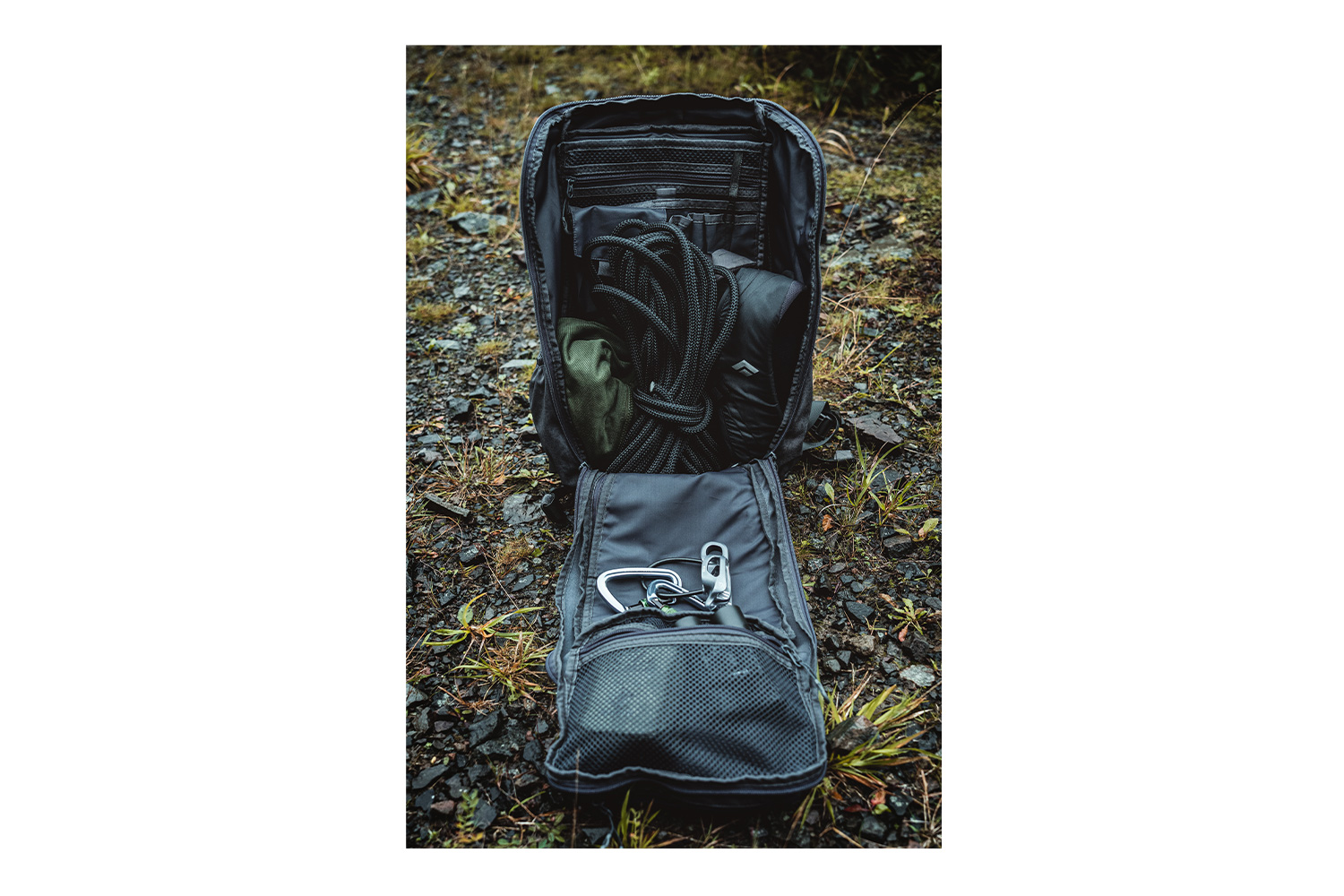 Offener schwarzer taktischer Rucksack auf felsigem Boden, gefüllt mit Kletterseil, Ausrüstungstaschen und Metallkarabinern im Inneren.