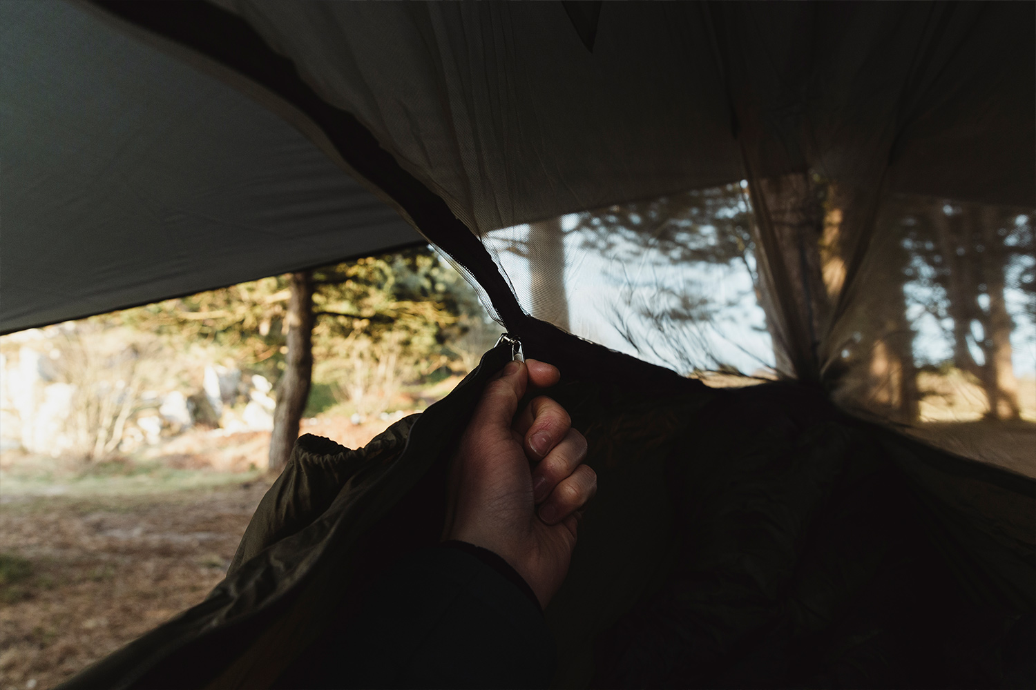 Eine Hand zieht den Reißverschluss an der Netztür eines olivgrünen Außenzeltes zu, im Hintergrund ist eine verschwommene Aussicht auf den Wald zu sehen.