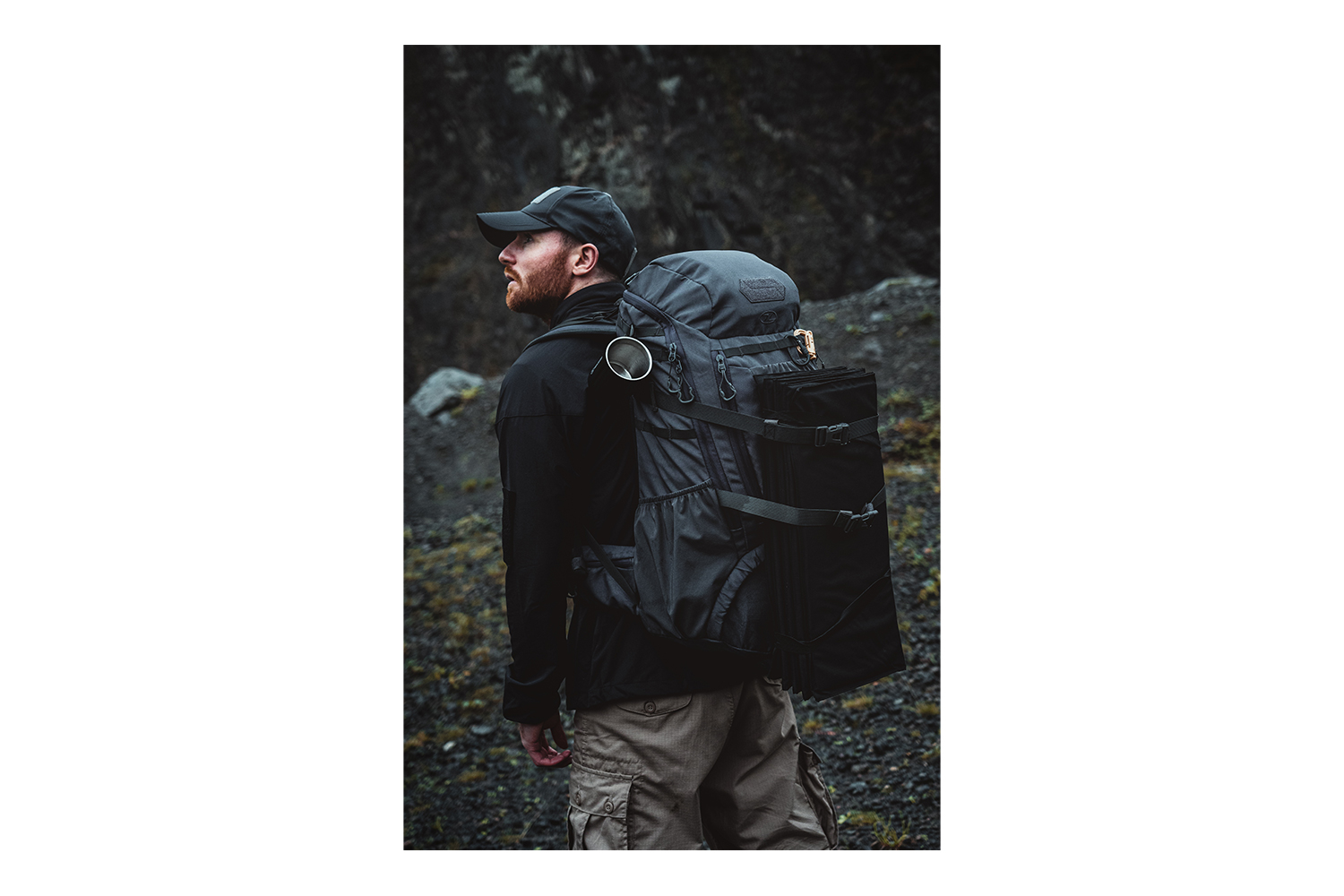 Ein Mann mit schwarzer Mütze und Jacke trägt einen dunkelgrauen taktischen Rucksack mit einer gefalteten Matte in einem rauen Außenbereich.
