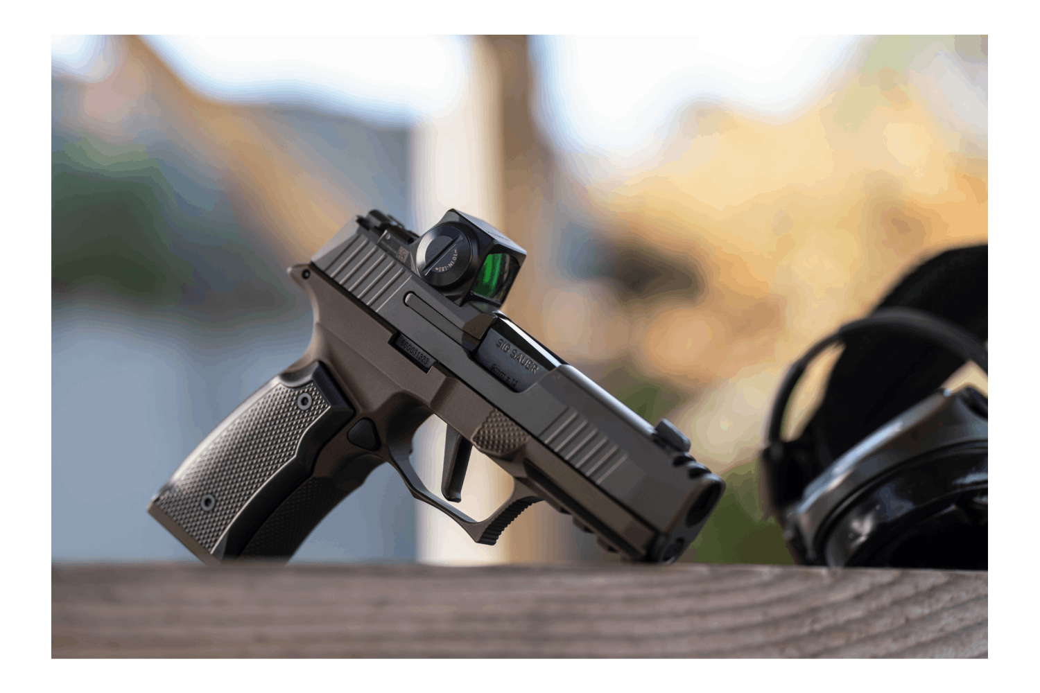 Schwarze Sig Sauer-Pistole mit Leuchtpunktvisier, auf einer Holzoberfläche ruhend, in der Nähe ein Gehörschutz.