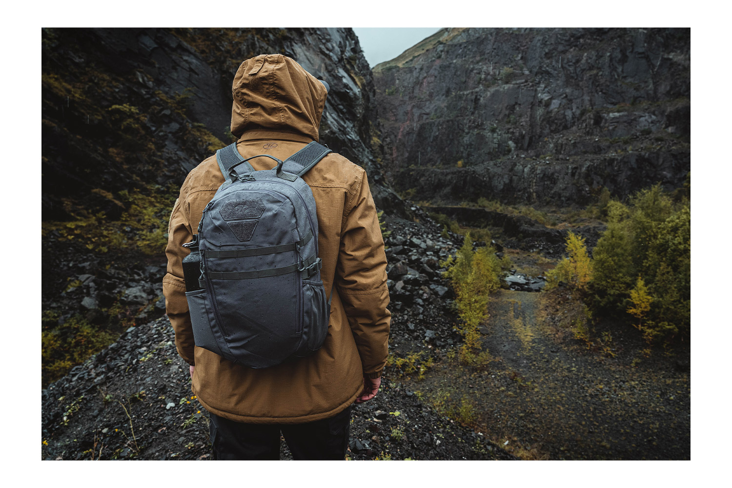 Eine Person, die eine braune Kapuzenjacke und einen grauen taktischen Rucksack trägt, steht in einer felsigen, bergigen Landschaft im Freien.