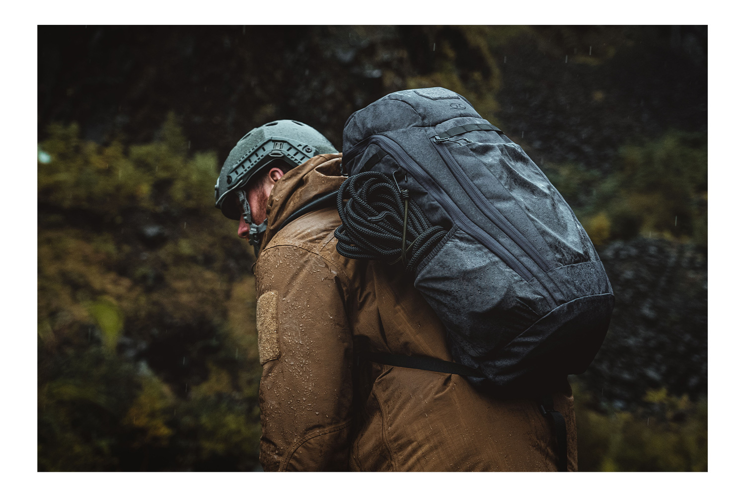Eine Person mit taktischem Helm, brauner Jacke und grauem Rucksack, die bei Regen ein schwarzes Seil trägt.