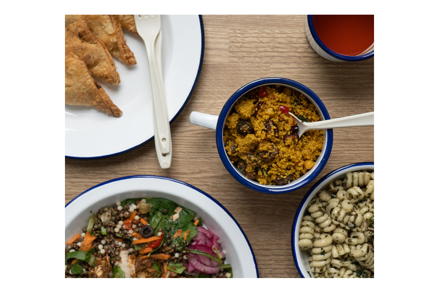 Draufsicht auf weiße Emailleschalen mit blauem Rand mit Couscous, Nudelsalat und Linsen auf einem Holztisch.
