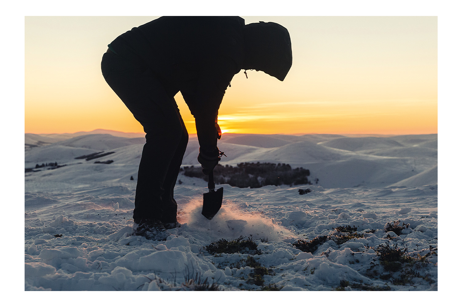 Die Silhouette einer Person, die während eines malerischen Wintersonnenuntergangs im Freien mit einer kompakten Klappschaufel im Schnee gräbt.