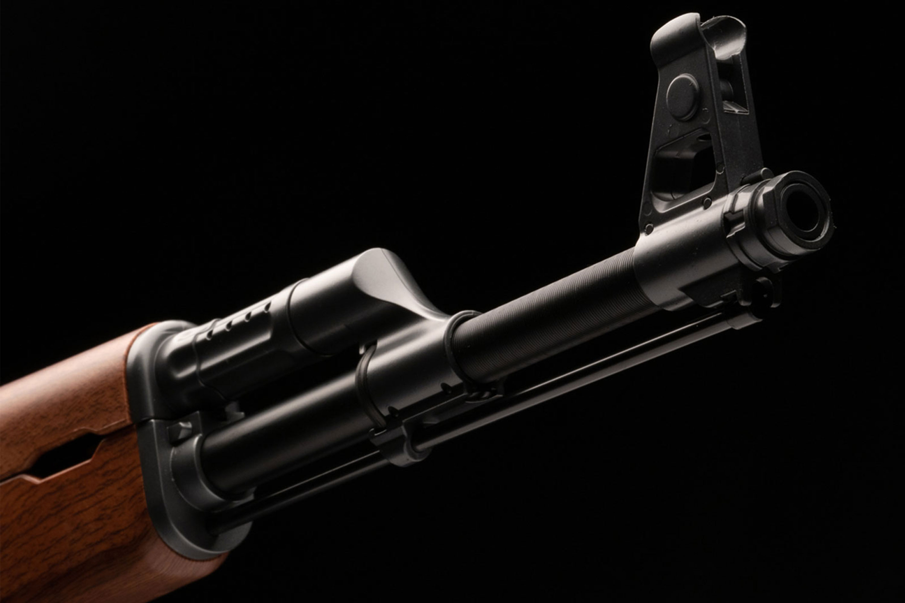 Nahaufnahme eines AK-47-Luftgewehrlaufs mit schwarzem Metallfinish und braunem Holzhandschutz auf dunklem Hintergrund.